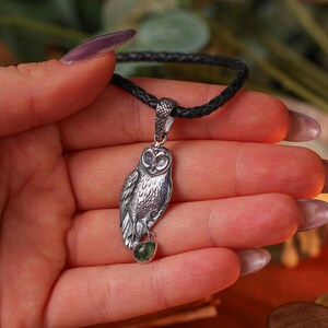 May include: A silver owl pendant on a dark braided cord. The owl pendant features detailed feathering and holds a small green gemstone. The pendant is held in a hand.