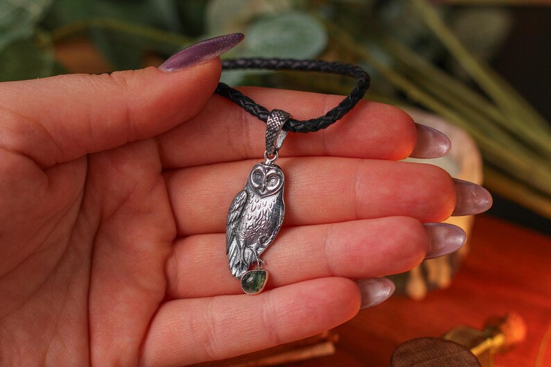 May include: A silver owl pendant on a dark braided cord. The owl pendant features detailed feathering and holds a small green gemstone. The pendant is held in a hand.