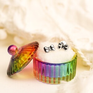 May include: A pair of silver bow-shaped earrings in a rainbow iridescent glass jewelry box with a white velvet lining.