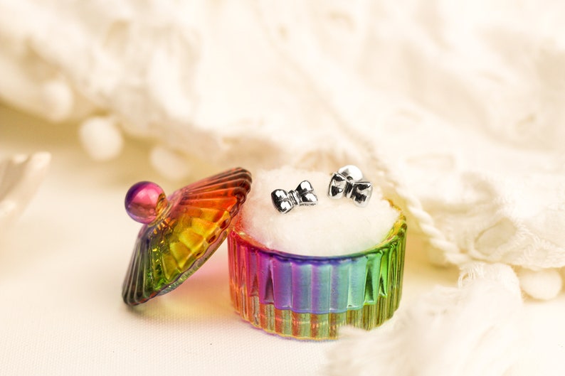 May include: A pair of silver bow-shaped earrings in a rainbow iridescent glass jewelry box with a white velvet lining.