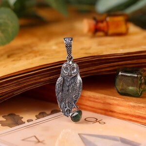 May include: A silver owl pendant with a green gemstone, displayed on a vintage book with a deck of cards. The owl pendant is detailed with intricate feather patterns. Small glass bottles are in the background.