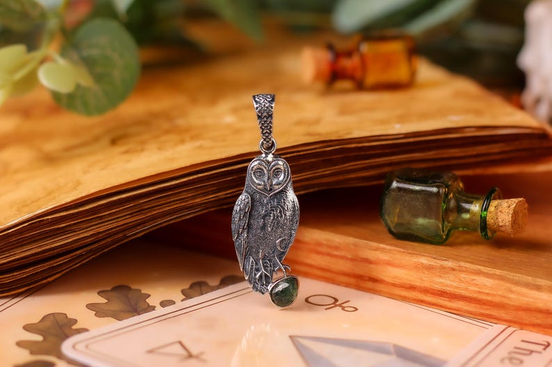 May include: A silver owl pendant with a green gemstone, displayed on a vintage book with a deck of cards. The owl pendant is detailed with intricate feather patterns. Small glass bottles are in the background.