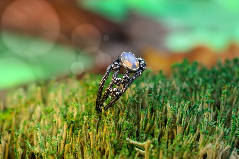 May include: A silver ring with a delicate floral design and a white opal gemstone. The ring is set on a bed of green moss.