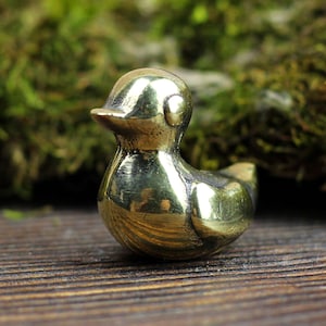Op de afbeelding: Een klein, glanzend, goudkleurig metalen vogelbeeldje. De vogel heeft een rond lichaam en een kleine snavel. Het staat op een houten oppervlak.