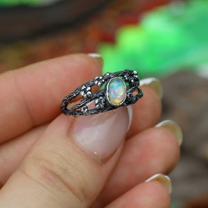 May include: A silver ring with a delicate, intricate design featuring a branch-like pattern with small flower accents. The ring has a white opal gemstone in the center.