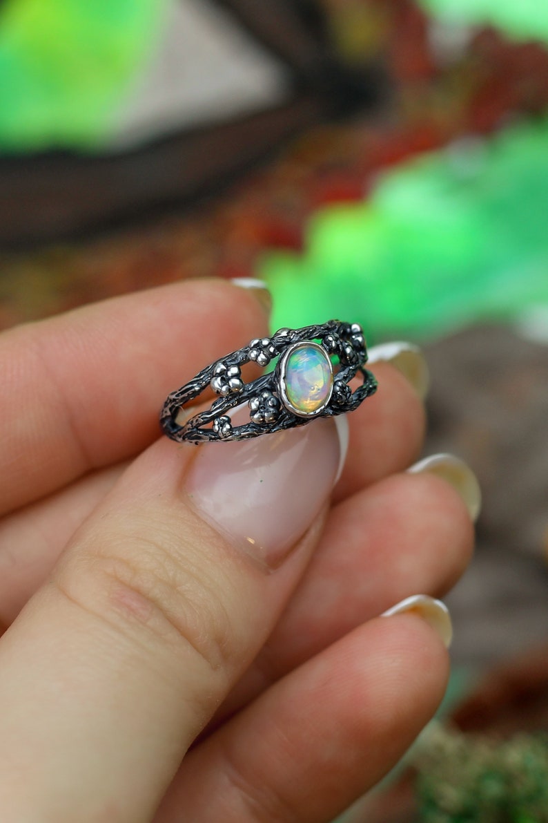 May include: A silver ring with a delicate, intricate design featuring a branch-like pattern with small flower accents. The ring has a white opal gemstone in the center.