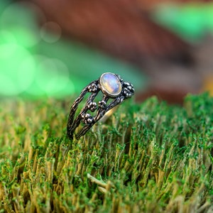 May include: A silver ring with a delicate floral design and a white opal gemstone. The ring is set against a backdrop of green moss.