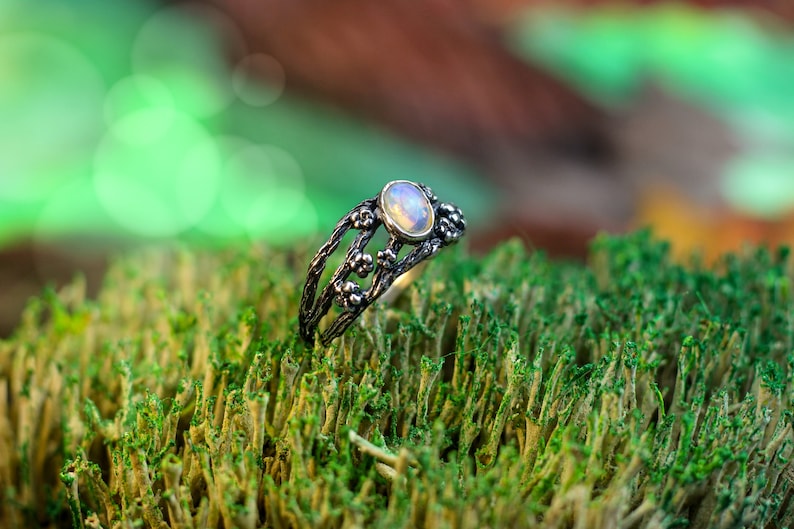 May include: A silver ring with a delicate floral design and a white opal gemstone. The ring is set against a backdrop of green moss.