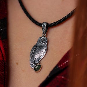 May include: A silver owl pendant with a green gemstone, hanging from a black braided cord necklace. The owl pendant features etched feathers and eyes. The necklace is worn against a red and black plaid shirt.