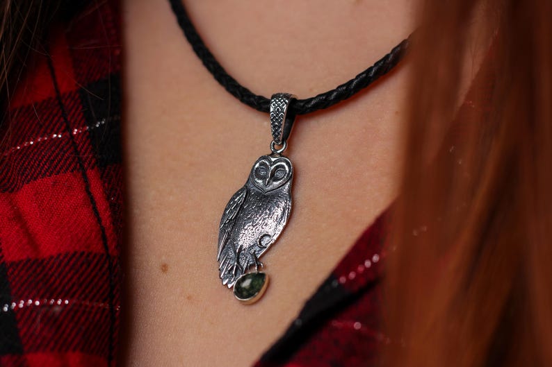 May include: A silver owl pendant with a green gemstone, hanging from a black braided cord necklace. The owl pendant features etched feathers and eyes. The necklace is worn against a red and black plaid shirt.