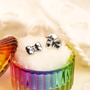 May include: A pair of silver bow-shaped earrings in a rainbow-colored glass jewelry box with a white cotton lining.