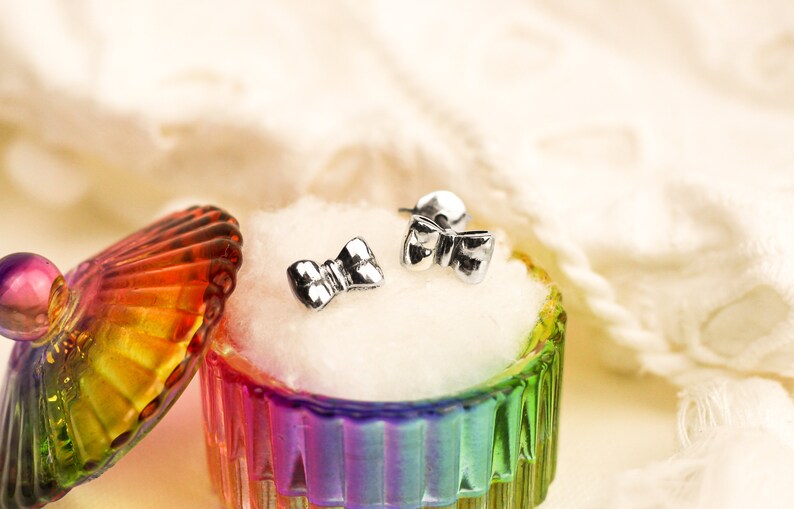 May include: A pair of silver bow-shaped earrings in a rainbow-colored glass jewelry box with a white cotton lining.