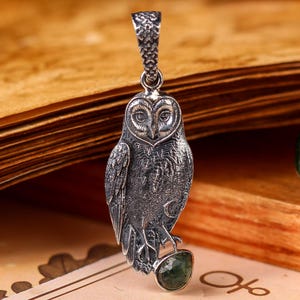 May include: A detailed silver owl pendant with a textured finish, perched on a green gemstone. The owl is suspended from a decorative bail. The pendant is displayed against an open book.