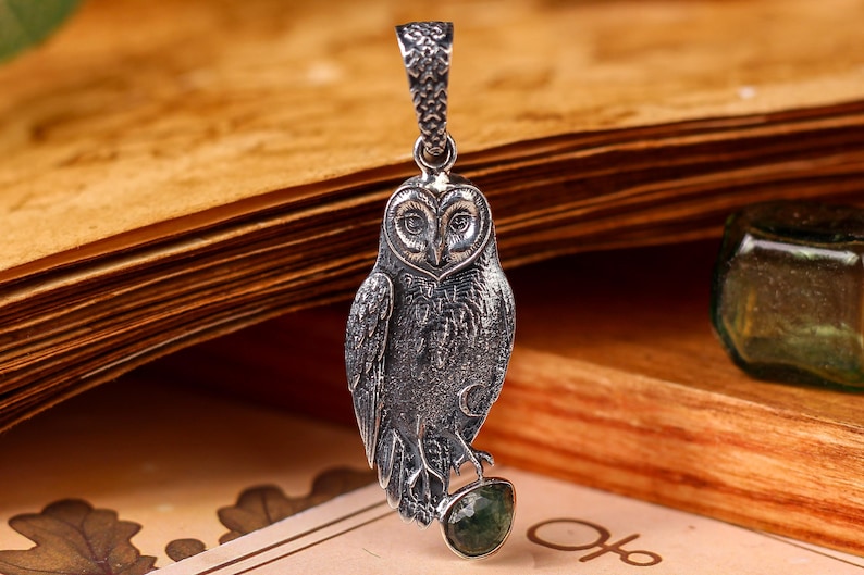 May include: A detailed silver owl pendant with a textured finish, perched on a green gemstone. The owl is suspended from a decorative bail. The pendant is displayed against an open book.