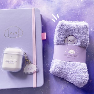 May include: A lavender notebook with a silver dog illustration and a clear case with a silver dog keychain. A pair of lavender fuzzy socks with a silver dog illustration and the text "cozy plush socks" on the tag.