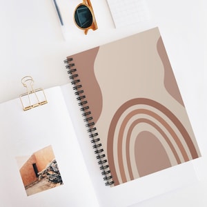 May include: A spiral bound notebook with a beige cover featuring a rainbow design in shades of brown. The rainbow is made up of five arches, with the largest arch at the bottom and the smallest arch at the top. The notebook is perfect for taking notes, journaling, or sketching.
