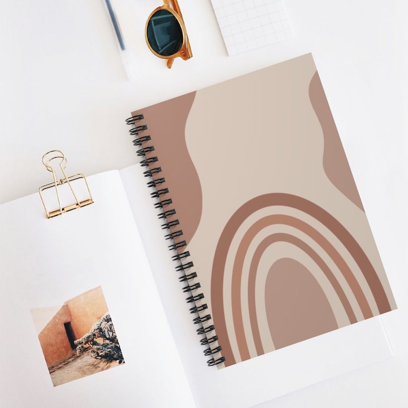 May include: A spiral bound notebook with a beige cover featuring a rainbow design in shades of brown. The rainbow is made up of five arches, with the largest arch at the bottom and the smallest arch at the top. The notebook is perfect for taking notes, journaling, or sketching.