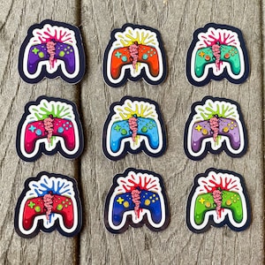 May include: Nine colorful video game controller stickers. Each sticker features a controller design with a brain-like graphic splitting the controller in half. The controllers are in various colors, including purple, red, blue, and green, with a black outline.