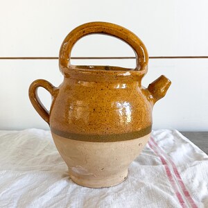 May include: A handcrafted, rustic ceramic pitcher with a rounded body and a large arched handle. The pitcher features a warm, earthy color palette with a speckled, glazed finish. It has a spout and a small handle on the side.
