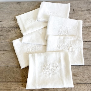 May include: A collection of white linen napkins, each embroidered with elegant monograms in a matching white thread. The napkins are neatly folded and arranged on a rustic wooden surface, showcasing their delicate texture and design.