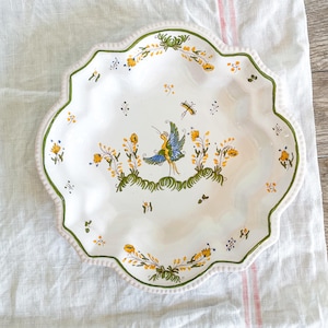 May include: White ceramic serving platter with a scalloped edge and a hand-painted design. The design features a central bird figure with yellow, blue, and green accents, surrounded by floral and insect motifs. The rim is trimmed with green and pink.