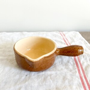 May include: A small, brown ceramic sauce bowl with a handle. The interior is a light beige color, and the exterior is a darker brown. The bowl is sitting on a white cloth with red stripes.