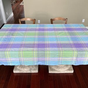 May include: A large rectangular tablecloth with a plaid pattern in shades of blue, green, and purple. The tablecloth is draped over a table, with the edges hanging down. The table is supported by two stone blocks.