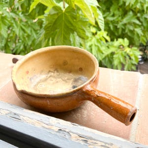 May include: A small, brown earthenware pot with a yellow interior and a single handle. The pot has a speckled glaze and appears to be vintage.