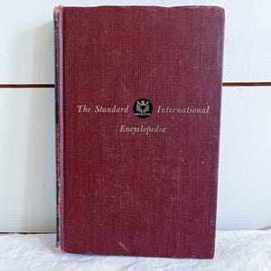 May include: A vintage burgundy-colored hardcover book titled "The Standard International Encyclopedia." The book has a textured cover with the title and a small emblem in gold lettering. The spine is a darker shade of burgundy.
