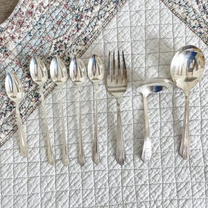 May include: An assortment of silver serving utensils, including spoons, forks, and a ladle, arranged on a white quilted surface. The silverware has a reflective surface and decorative handle details. The background includes a floral patterned fabric.