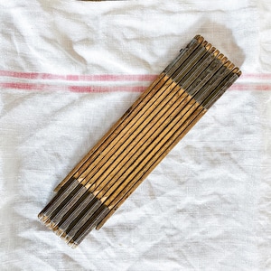 May include: A vintage wooden folding ruler with metal end caps, resting on a white cloth with a red stripe. The ruler is made of multiple wooden segments joined together, creating a zig-zag pattern when folded. The wood has a warm, aged tone.