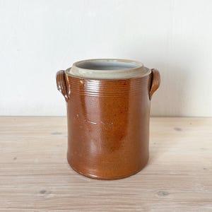May include: A brown stoneware crock with a lid, featuring two side handles. The cylindrical container has a glossy finish and a light-colored rim. The crock is likely used for food storage or decoration. The background is a light neutral color.