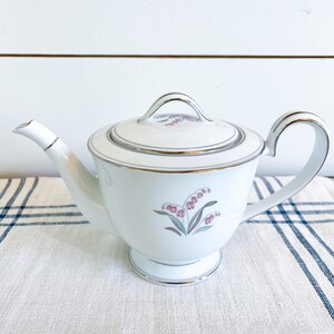 May include: White porcelain teapot with a lid, handle, and spout. The teapot features a floral design in pink and gray, with silver trim around the edges. The teapot is sitting on a white and blue striped cloth.