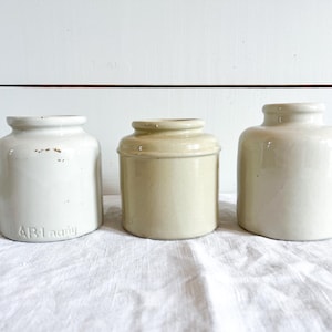 May include: Three antique ceramic jars in varying shades of cream and beige. The jar on the left has the text "AR-I agny" printed on it. The jars are displayed on a white cloth.