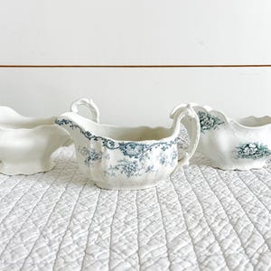 May include: Three white ceramic gravy boats. One is plain, the second has a blue floral pattern, and the third has a green floral pattern. All have handles and decorative edges. They are arranged on a white quilted surface.