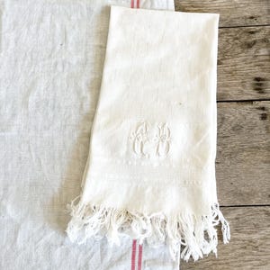 May include: White linen towel with a checkered pattern and embroidered initials. The towel has a fringed edge and is placed on a white linen cloth with red stripes. The background is a wooden surface.