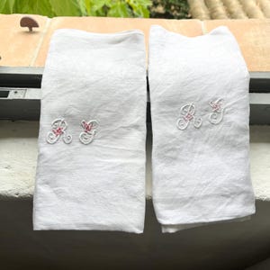 May include: Two white linen tea towels with pink embroidered flowers and the letter "R" on each towel.