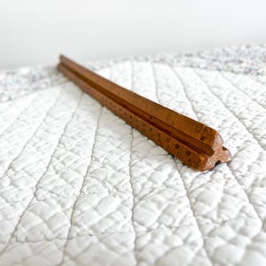 May include: A vintage wooden ruler with etched markings, likely used for measuring. The ruler is a warm brown color and appears to be well-worn. It is resting on a white quilted surface.