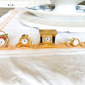 May include: A collection of miniature gold-colored clocks, including an alarm clock, a mantle clock, a classical-style clock, and an airplane-shaped clock. The clocks are displayed on a white surface with an orange border.