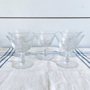 May include: Set of clear, etched glass champagne coupe glasses. Each glass features a wide bowl, a short stem, and a round base. The glasses are arranged on a white and blue striped cloth.