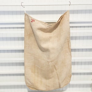 May include: A large, off-white linen sack with the initials "JP" embroidered in red thread. The sack is hanging from two hooks on a white wall.