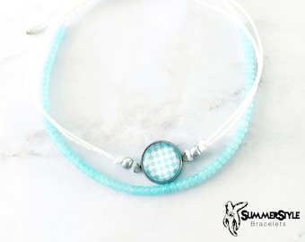May include: A white and light blue beaded bracelet with a checkered charm. The bracelet is tied with a knot and has a silver bead.