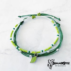 Op de afbeelding: Een groene en witte kralen armband met een groene bladeren bedel. De armband is gemaakt van touw en heeft een zilveren accent. De armband ligt op een wit marmeren oppervlak. SummerStyle Bracelets.
