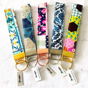 May include: Five fabric wristlet keychains with diverse patterns including floral, geometric, and patchwork designs. Each features a metal clasp and a split ring, with small white rectangular tags attached that read SummerStyle Bracelets.