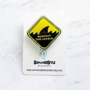 May include: Yellow diamond-shaped sign with a black shark fin and waves. The sign reads "Respect the Locals". The sign is on a white card with the text "SummerStyle Bracelets" and a website address.