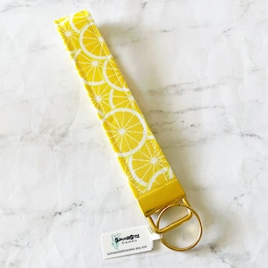 May include: Yellow and white lemon slice patterned wristlet key fob with a gold-colored metal ring and clasp. The key fob is made of fabric and has a tag that says "SummerStyle Bracelets" and the Etsy shop URL.