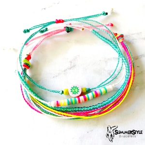 以下が含まれることがあります： 太陽のチャームが付いたカラフルな友情ブレスレットのセット。ブレスレットは、ピンク、グリーン、イエロー、ホワイトなど、さまざまな色の糸とビーズで作られています。