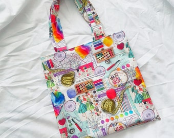 Pode incluir: Uma bolsa tote branca com um design colorido que apresenta suprimentos de costura, linha, agulhas e tecido. A bolsa tem um fundo branco com detalhes coloridos e uma variedade de itens relacionados à costura.