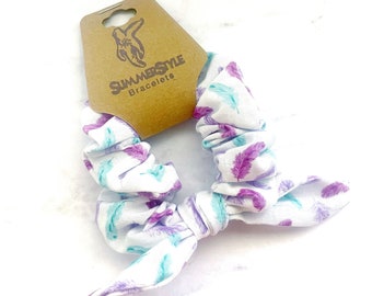 May include: White scrunchie with a purple and teal feather print. The scrunchie is tied in a bow and has a brown tag that says "SummerStyle Bracelets".
