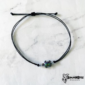May include: A black string bracelet with a small iridescent turtle charm and white beads.